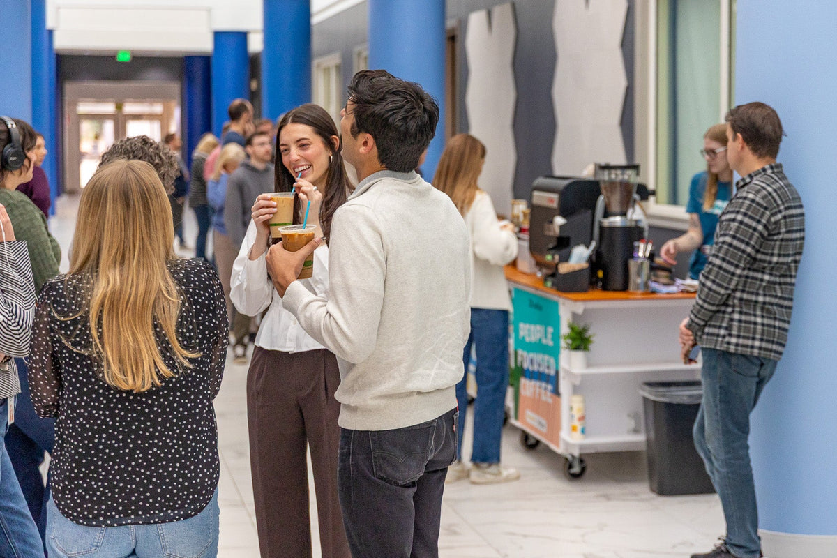 A group of people gather around the 321 Coffee Cart at an event and enjoy their coffee while talking and laughing together.