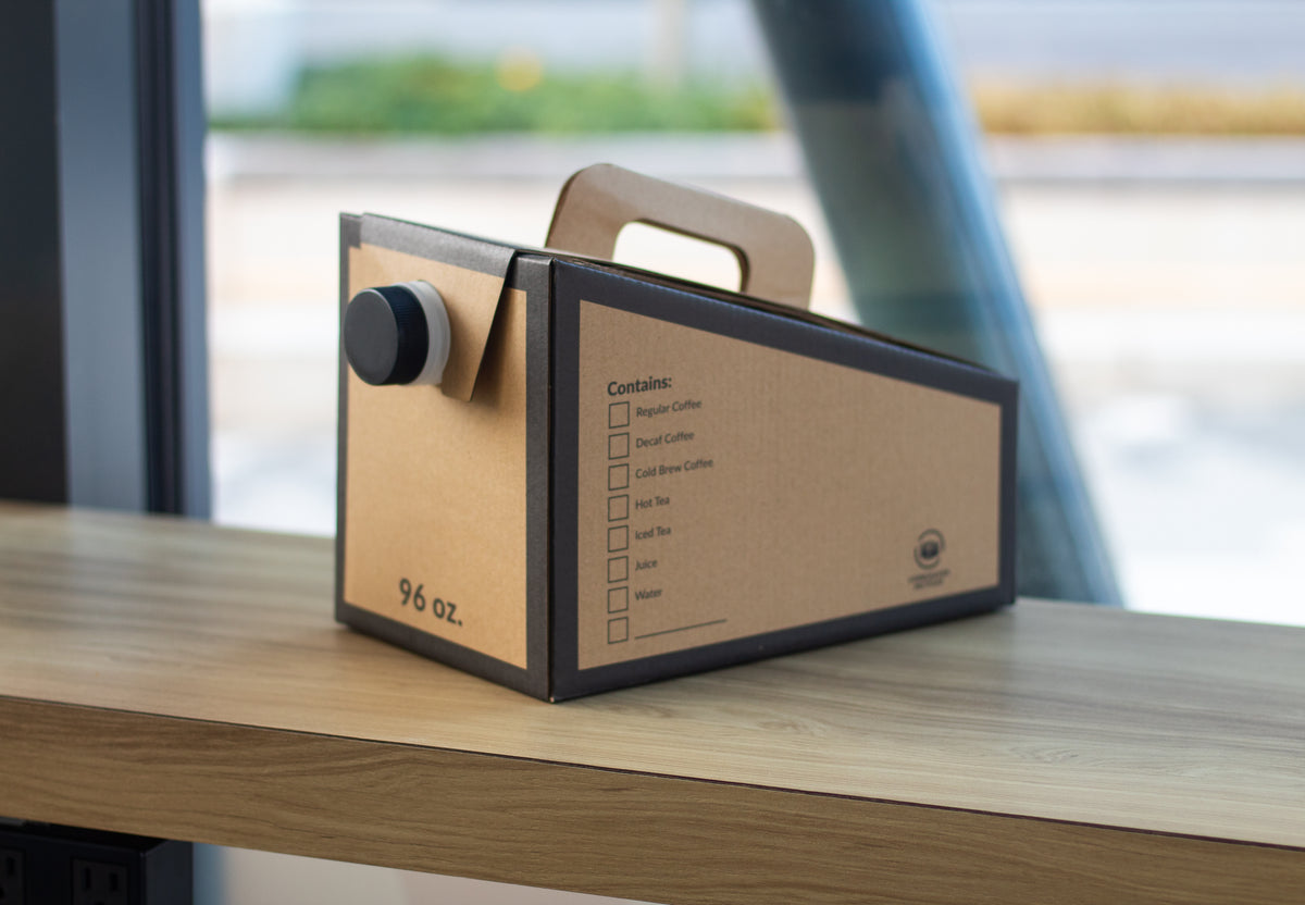 A disposable, cardboard 96 oz catering traveler sits on a wooden counter.