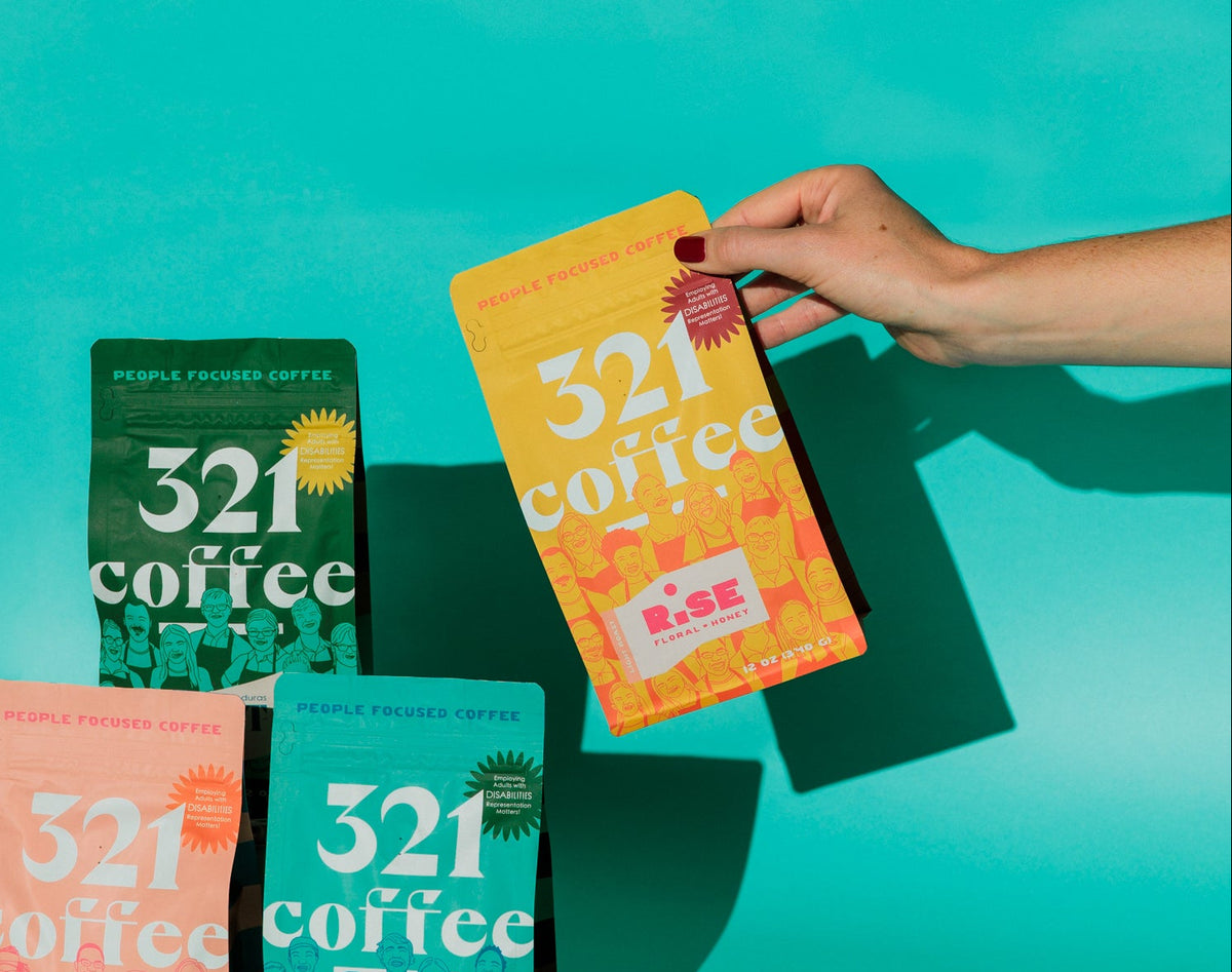A hand reaches in to pick up a yellow bag of Rise light roast coffee from a line up of 321 Coffee's core roasts on a teal background.
