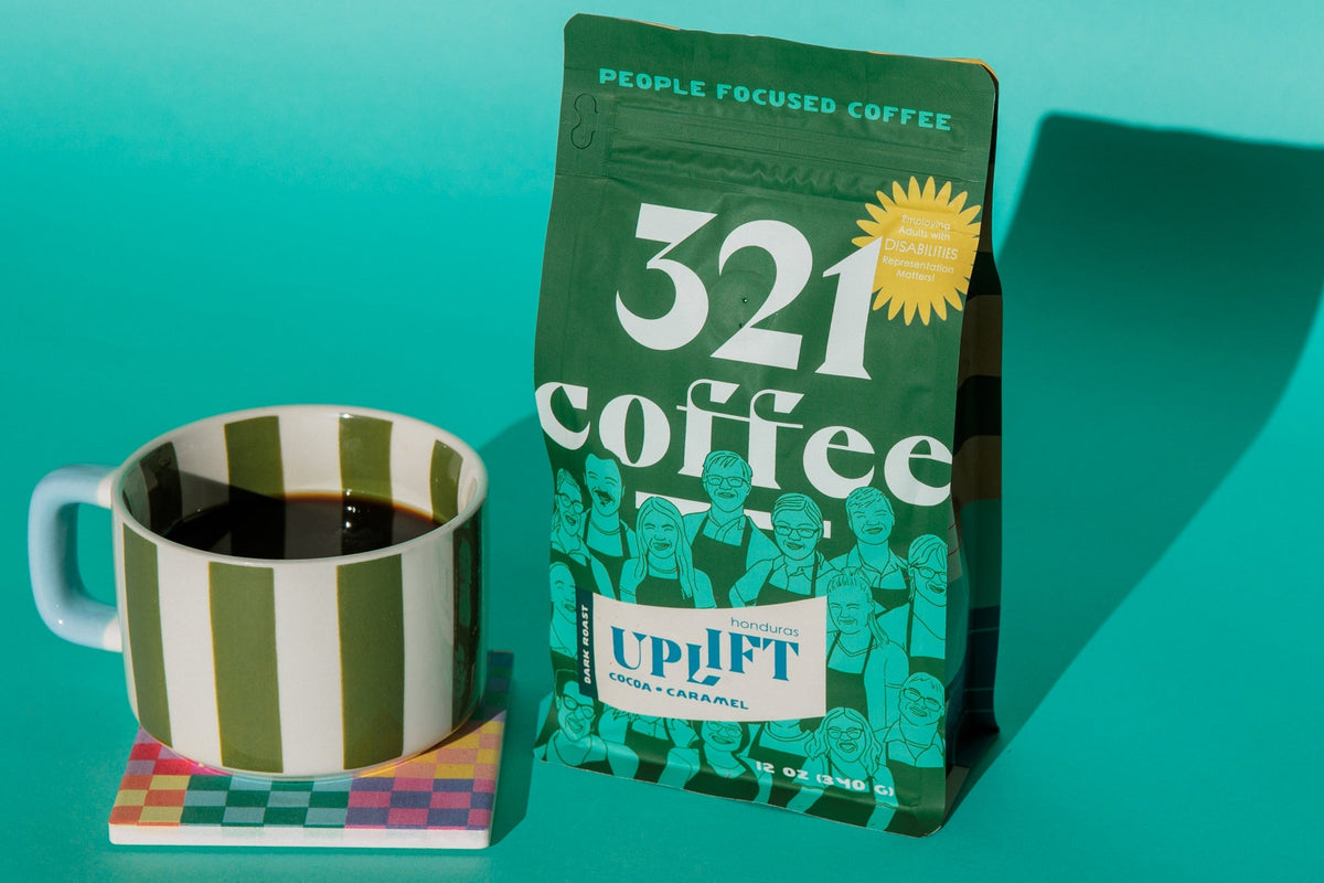 A green bag of Uplift, 321 Coffee's dark roast, sits next to a green and white striped hot coffee mug. Both are placed on a teal background.