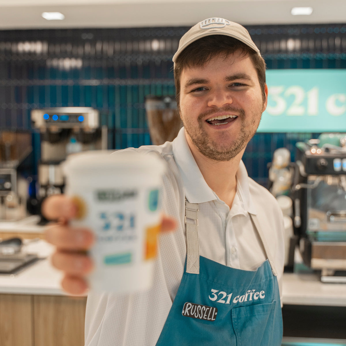 A barista wearing the signature blue 321 Coffee apron stands behind the counter and holds out a 321 Coffee hot cup to the camera.