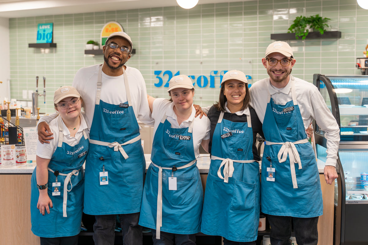 Five baristas with developmental disabilities stand together and smile with their arms around each other in front of a counter in a 321 Coffee shop. They are all wearing a blue apron with a 321 Coffee logo, and a tan hat.