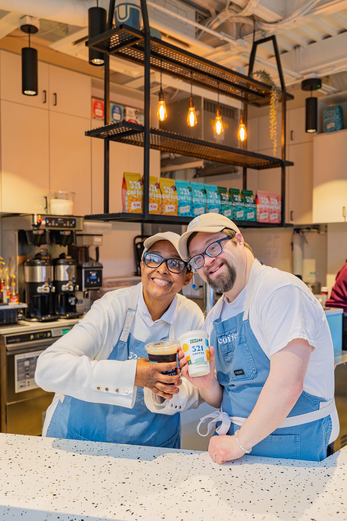 Two baristas with developmental disabilities stand together behind a counter in 321 Coffee's Downtown Raleigh shop and each hold a cup of coffee. They are wearing blue aprons with a 321 Coffee logo.