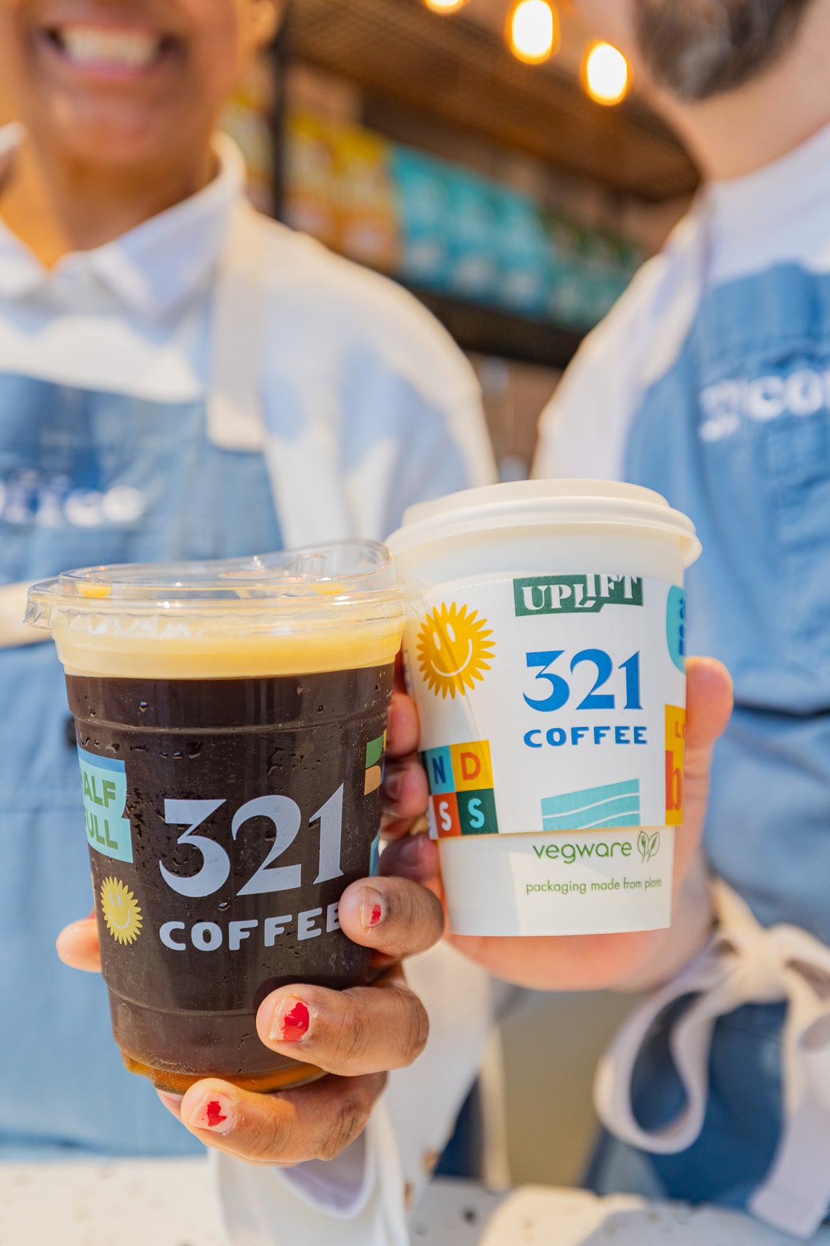 A cup of nitro cold brew and a cup of hot drip coffee are held together by two 321 Coffee baristas. The cups feature colorful brand icons with uplifting messages like "half full", "love big", and "kindness."