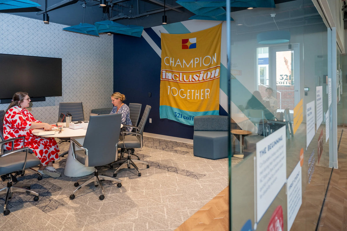 Two women work at a large round white conference room table in a conference room in the NC State shop.