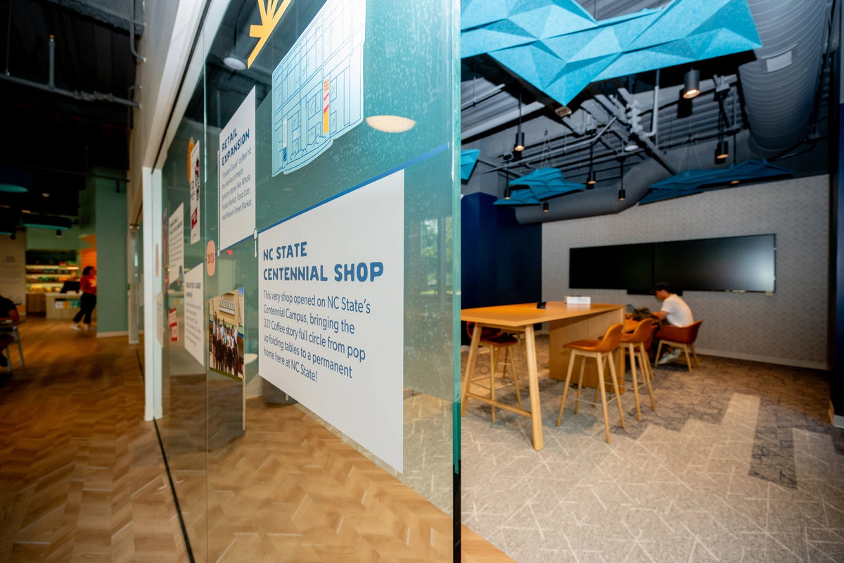 A spacious conference room in the NC Sate shop includes a large wooden rectangle table with leather chairs and a double tv display.