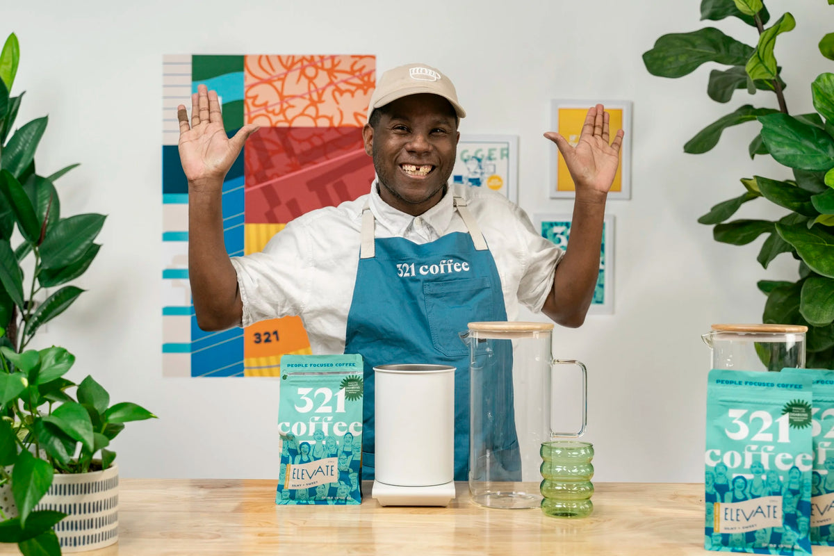 A 321 Coffee barista raises both hands in the air in excitement as he gets ready to start walking through a brew guide. He stands in front of a wall with colorful 321 Coffee branded signage.