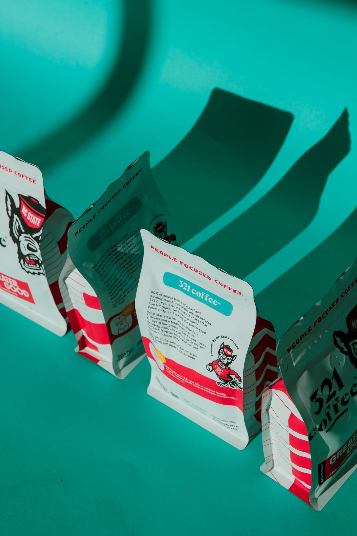 Four bags of Greater Good, 321 Coffee's licensed coffee with North Carolina State University, are displayed on a teal background. The bags are white with red accents, and feature the Mr. Wuf icon on the front.
