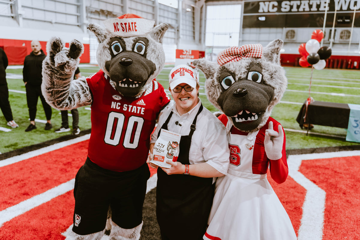 Sam holds a bag of Greater Good and stands in between Mr. and Ms. Wuf, the NC State Wolfpack mascots.