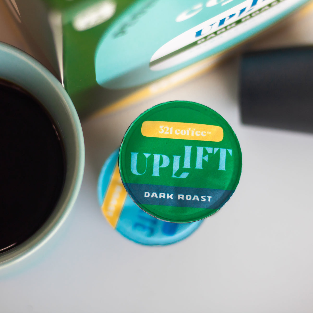 A green dark roast coffee pod is stacked on top of a teal medium roast coffee pod, sitting next to a box of coffee pods and a teal mug.