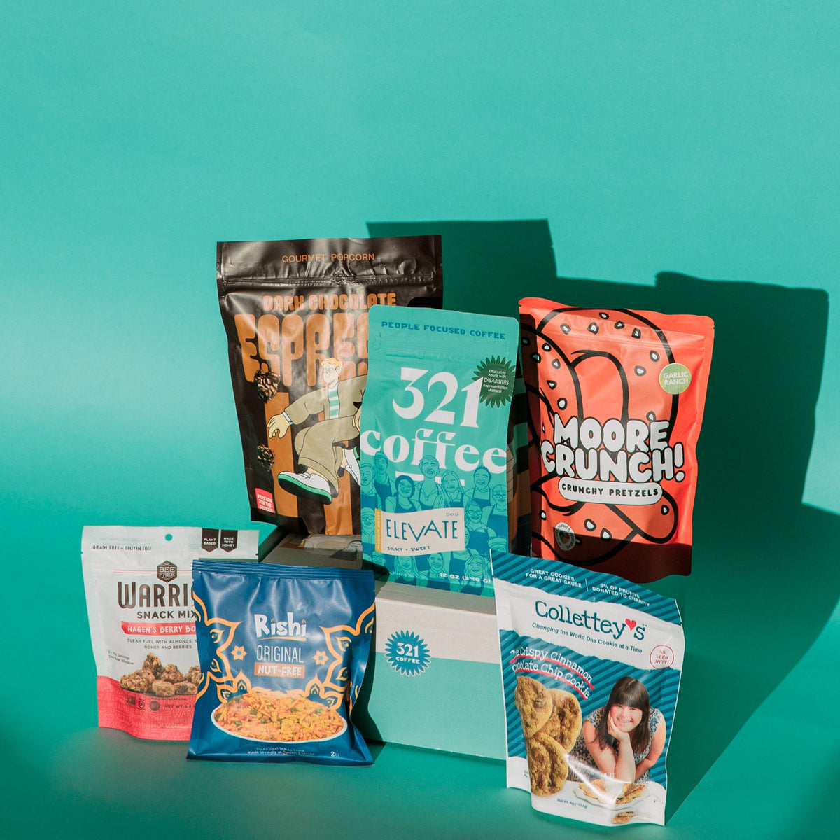 An assortment of snacks from inclusive companies is displayed on a teal background. In addition to 321 Coffee, the assortment includes Moore Crunch pretzels, Popcorn for the People, Warrior Snack mix, Rishi snacks, and Collettey's cookies.